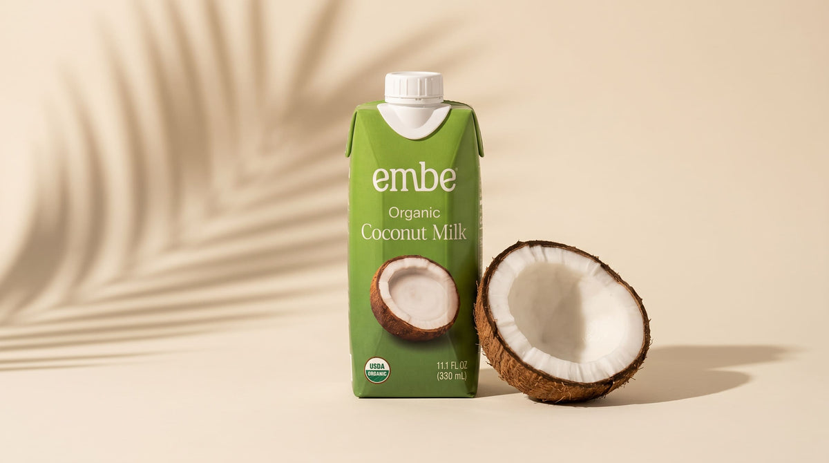 A green carton of embe Organic Coconut Milk with a white cap, standing on a beige surface next to a halved coconut. The background has soft, warm shadows.